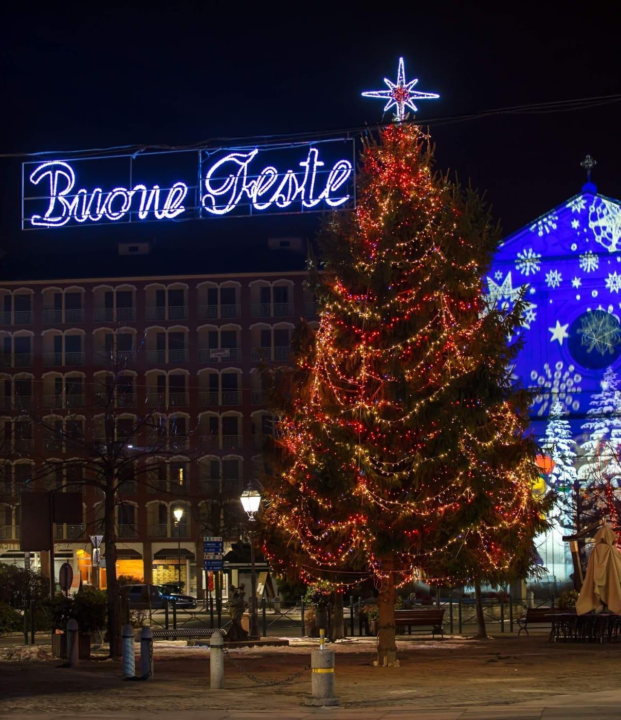 Scritta luminosa Buone Feste sospesa sopra una piazza, con albero e facciata illuminata