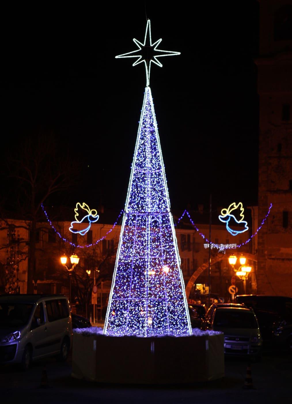 Cono luminoso azzurro con due angeli sospesi ai lati, in una piazza
